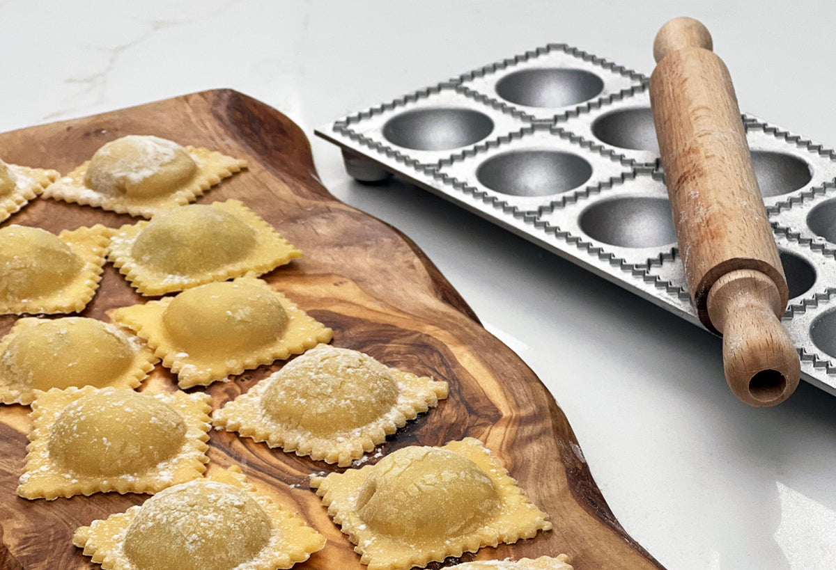 Uncooked ravioli on wooden board with metal mold and wooden rolling pin on white surface.