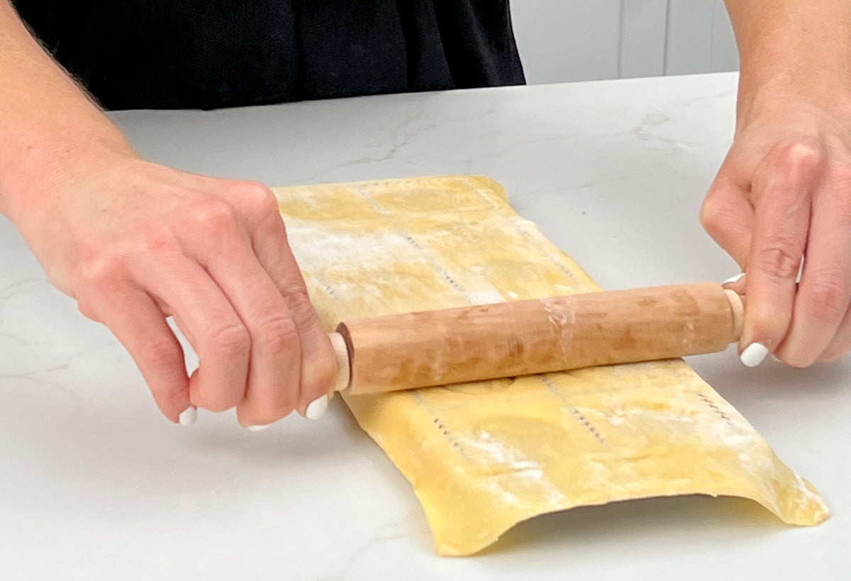 Hands rolling pasta dough with wooden rolling pin on marble surface.