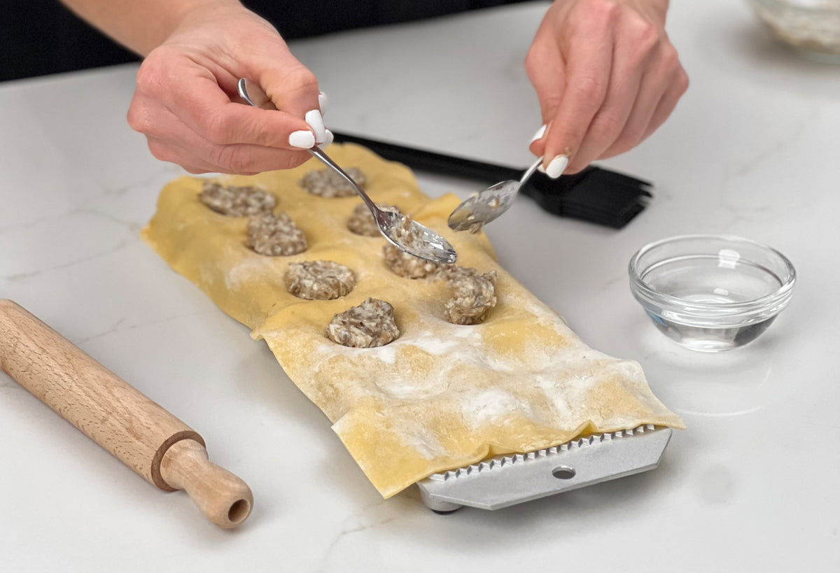 Hands filling pasta sheets with meat filling using two spoons, with rolling pin and small bowl nearby.