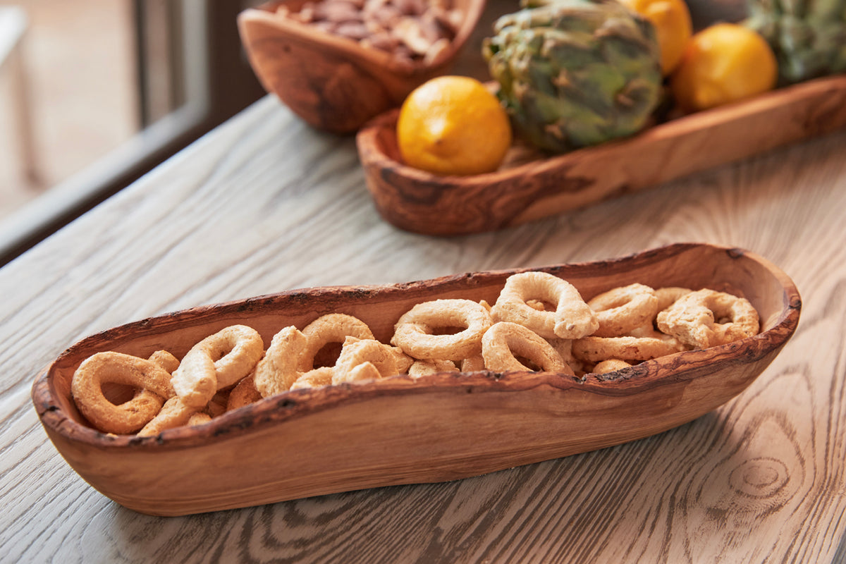 Wooden boat-shaped bowl with live edge containing snacks, displayed on white wood surface with fruits in background.