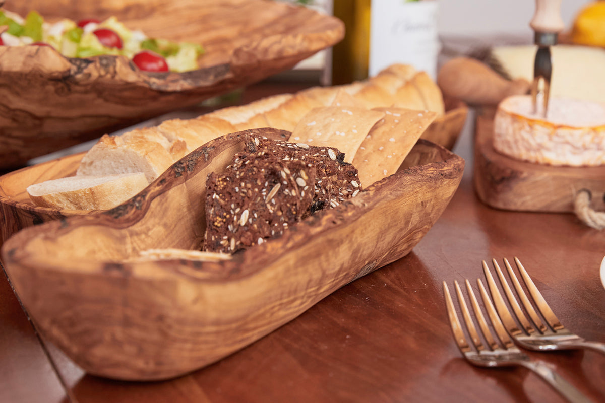 Wooden boat-shaped bowl with live edge holding bread and seeded roll on dining table.