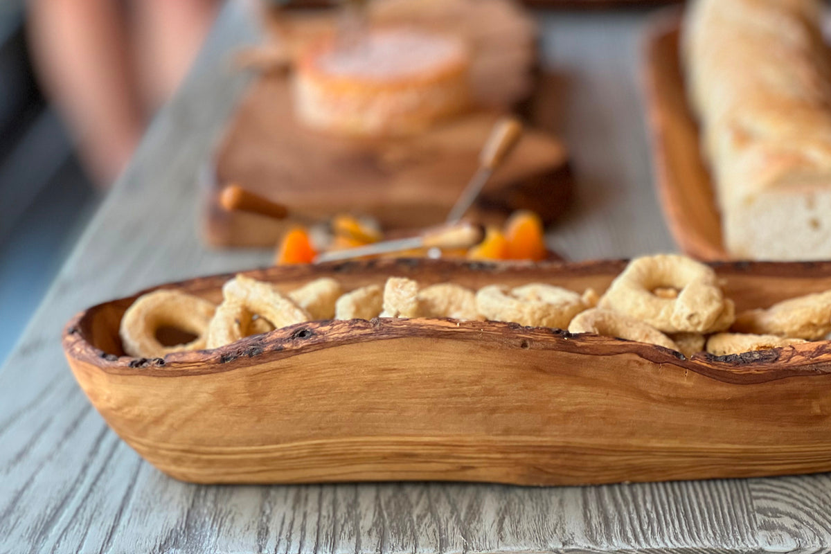 Wooden boat-shaped bowl with natural live edge containing crackers and breadsticks on a gray table.