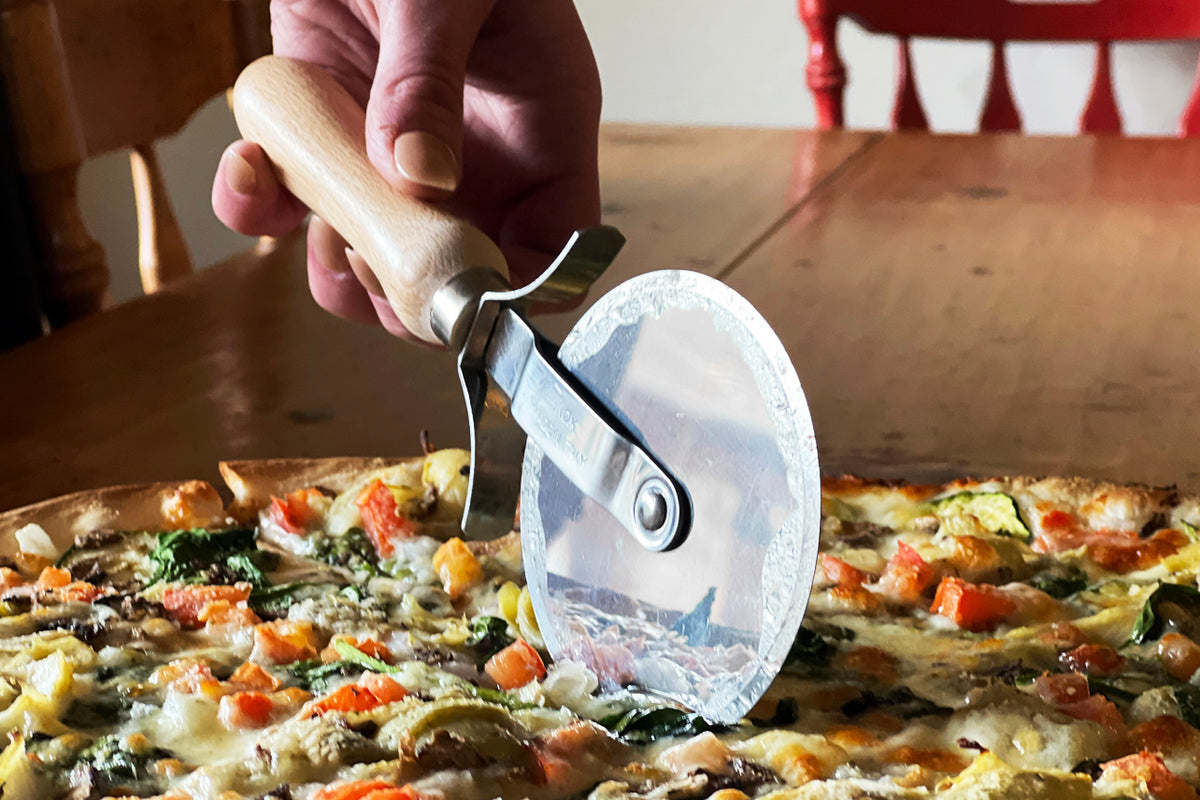 Hand holding wooden-handled pizza cutter with stainless steel wheel slicing into pizza topped with vegetables and cheese.