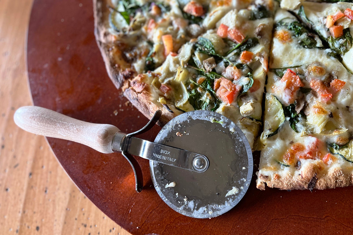 Circular pizza cutter with wooden handle resting on a sliced pizza with vegetables and toppings.