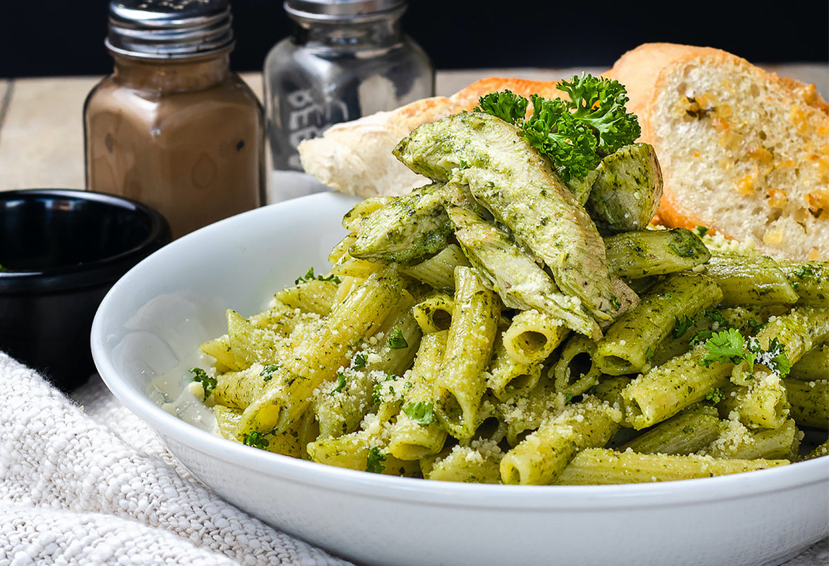 Penne pasta coated in green pesto sauce, garnished with parmesan and fresh basil, served with bread.