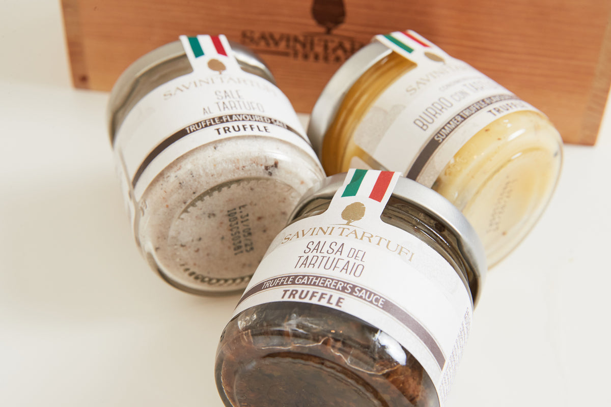 Three glass jars of Savini Tartufi truffle products with white labels and Italian flag ribbons, displayed with wooden box.