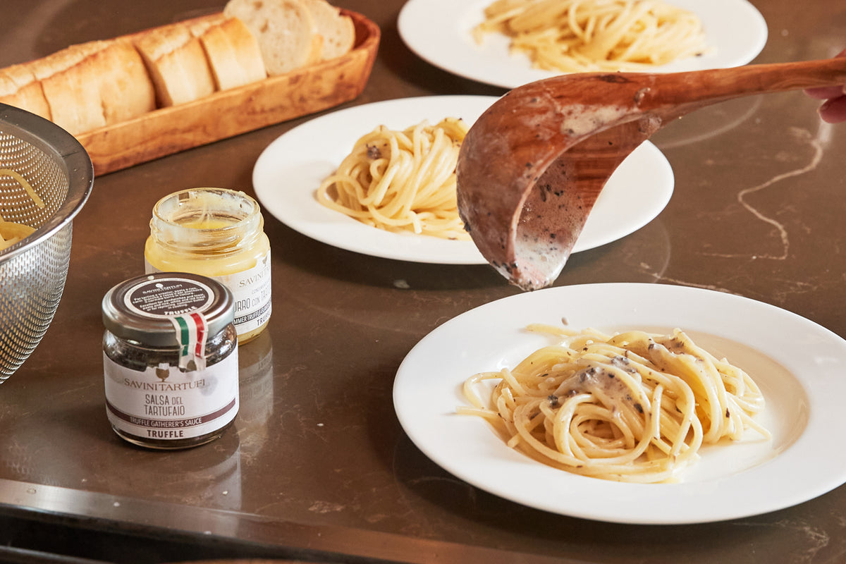 Glass jar of Savini Tartufi truffle sauce displayed with pasta dishes, bread, and cured meat on a brown table.