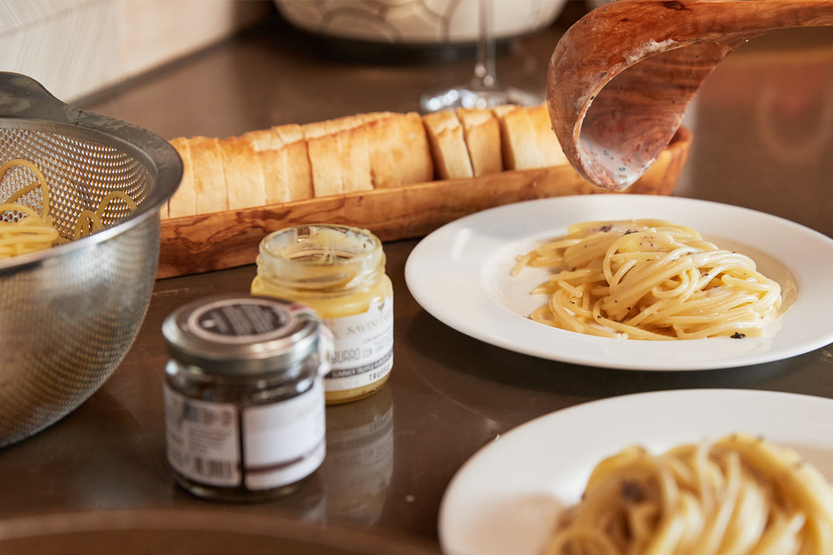 Truffle sauce jar displayed with pasta dishes, bread, sliced cured meat, and kitchen utensils on a wooden surface.