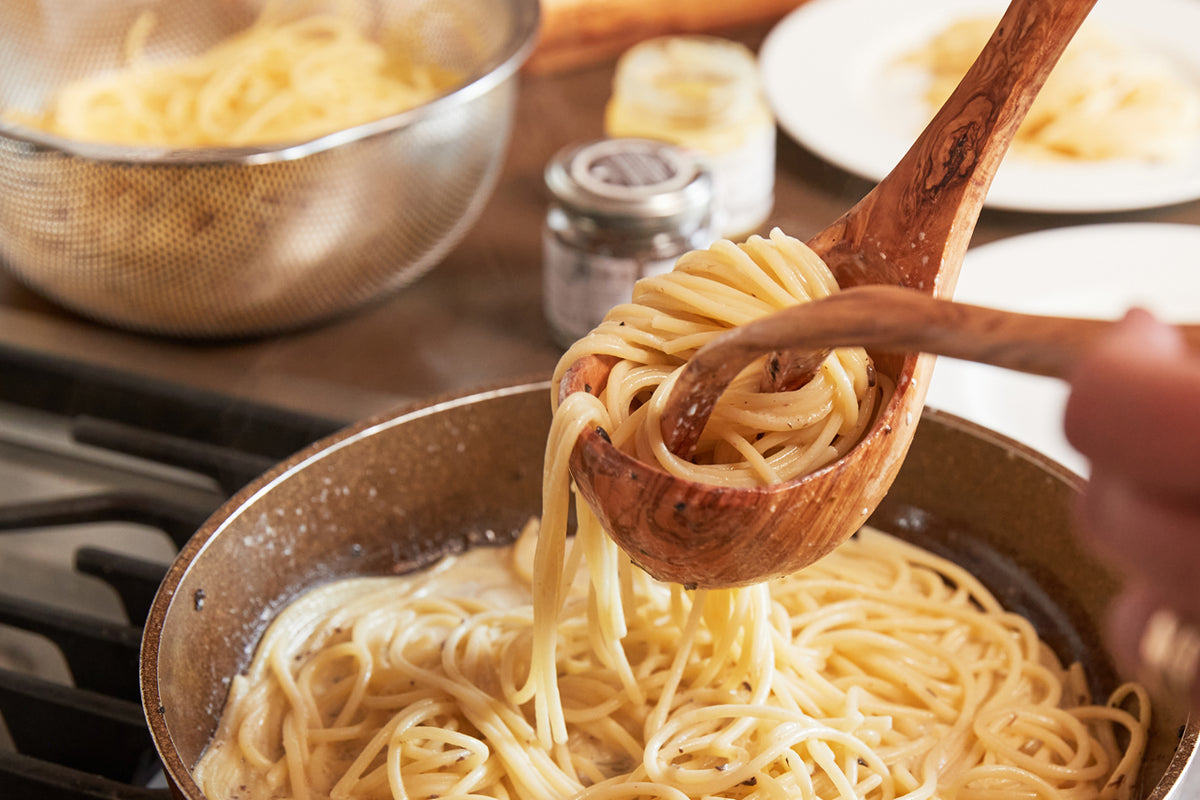 Wooden spoon lifting cooked spaghetti from a pan with pasta cooking in background.