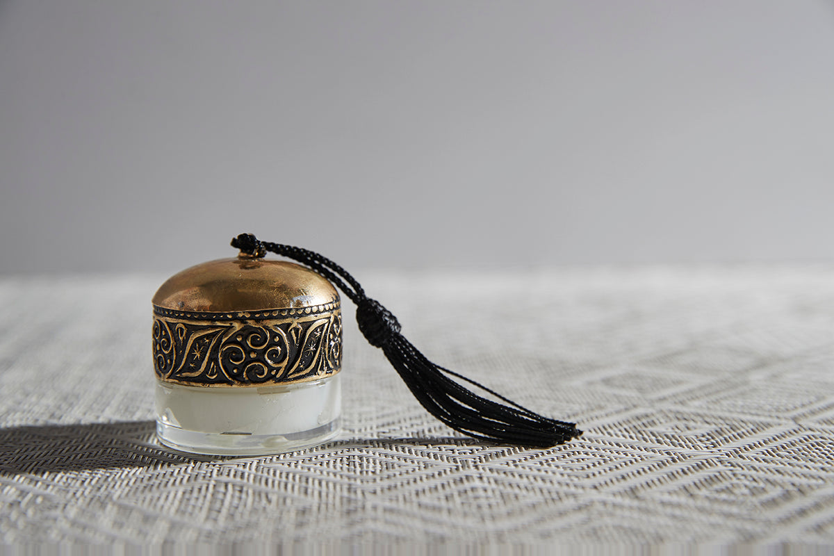 Small decorative lip balm container with gold filigree band, cream-colored balm visible, black tassel attached.