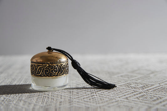 Small decorative lip balm container with gold filigree band, cream-colored balm visible, black tassel attached.