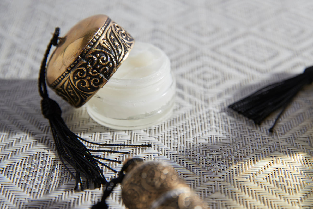 Small jar of white lip balm with ornate brass-toned lid and black tassel, on patterned fabric.