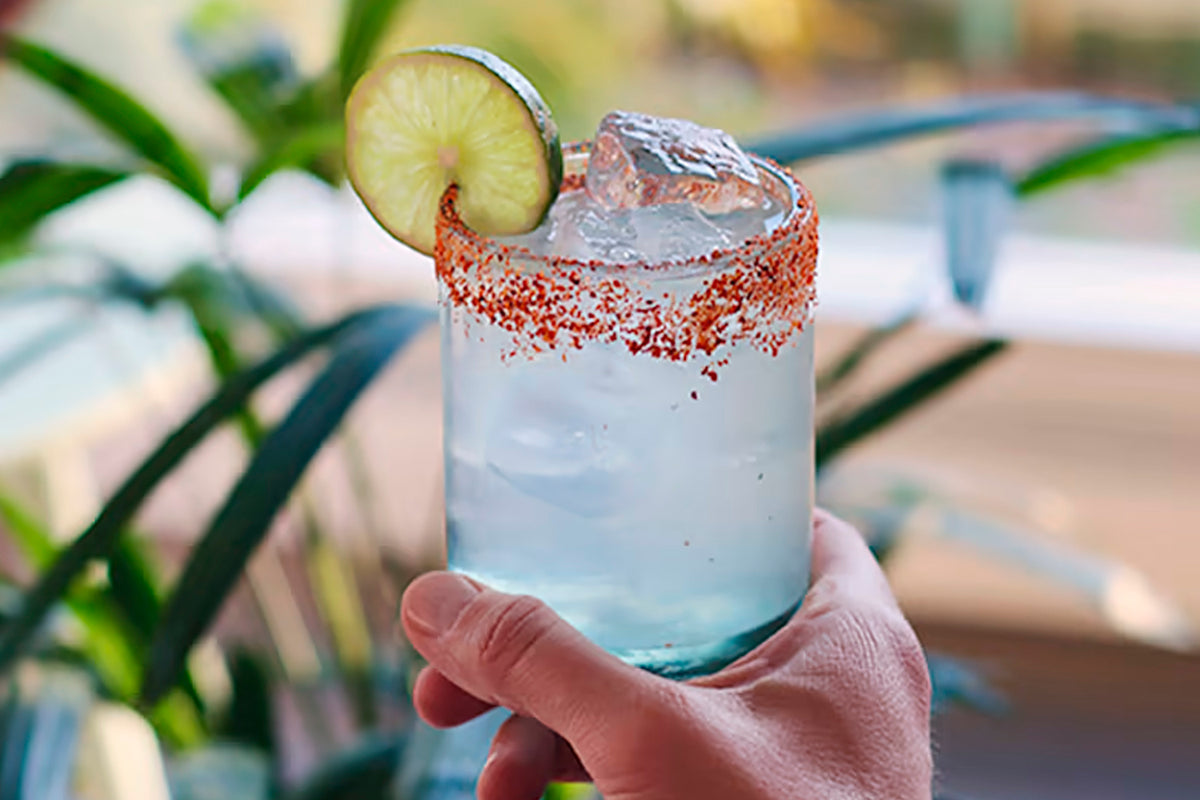 Cocktail with lime slice garnish, red-rimmed glass filled with clear liquid and ice, held in hand outdoors.