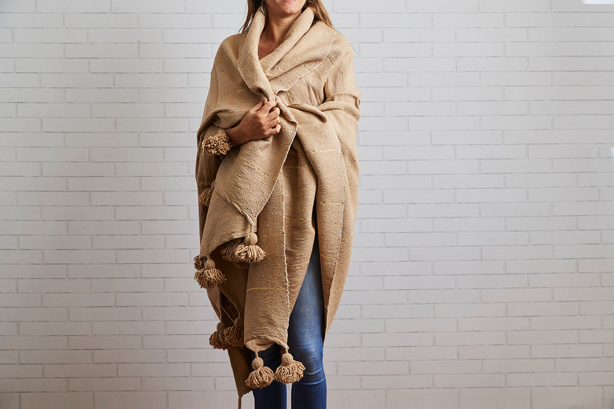 Woman wearing beige woven blanket with brown tassels draped over shoulders against white brick background.