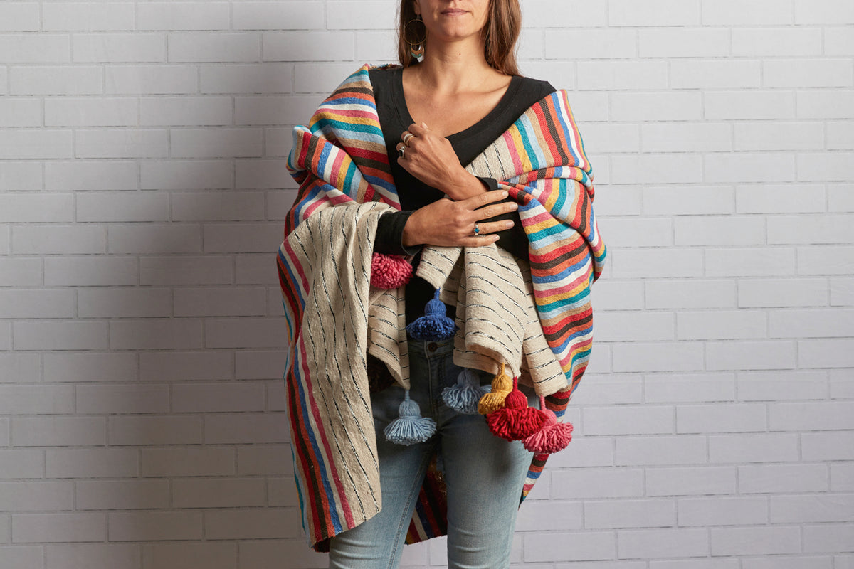 Woman wearing striped blanket with rainbow colors and tassels in red, blue, and yellow draped over shoulders.