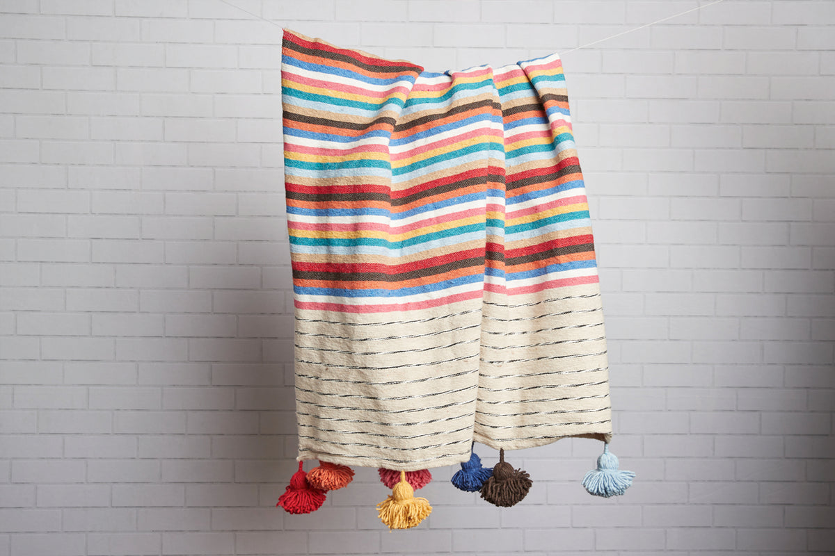 Woven blanket with horizontal stripes in red, blue, green, and cream, featuring colorful tassels at bottom edges.
