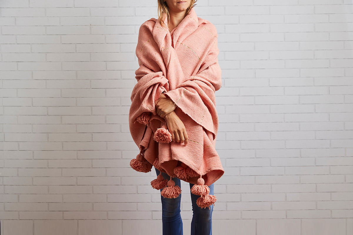 Terracotta wool blanket with tasseled edges draped around a person wearing jeans against a white brick wall.