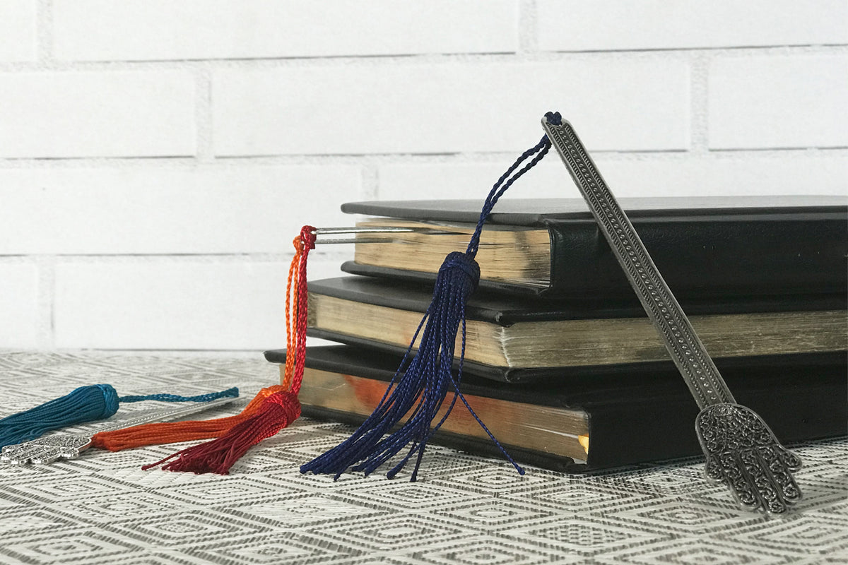 Three metal hand-shaped bookmarks with tassels in red, navy blue, and silver displayed with stacked books.