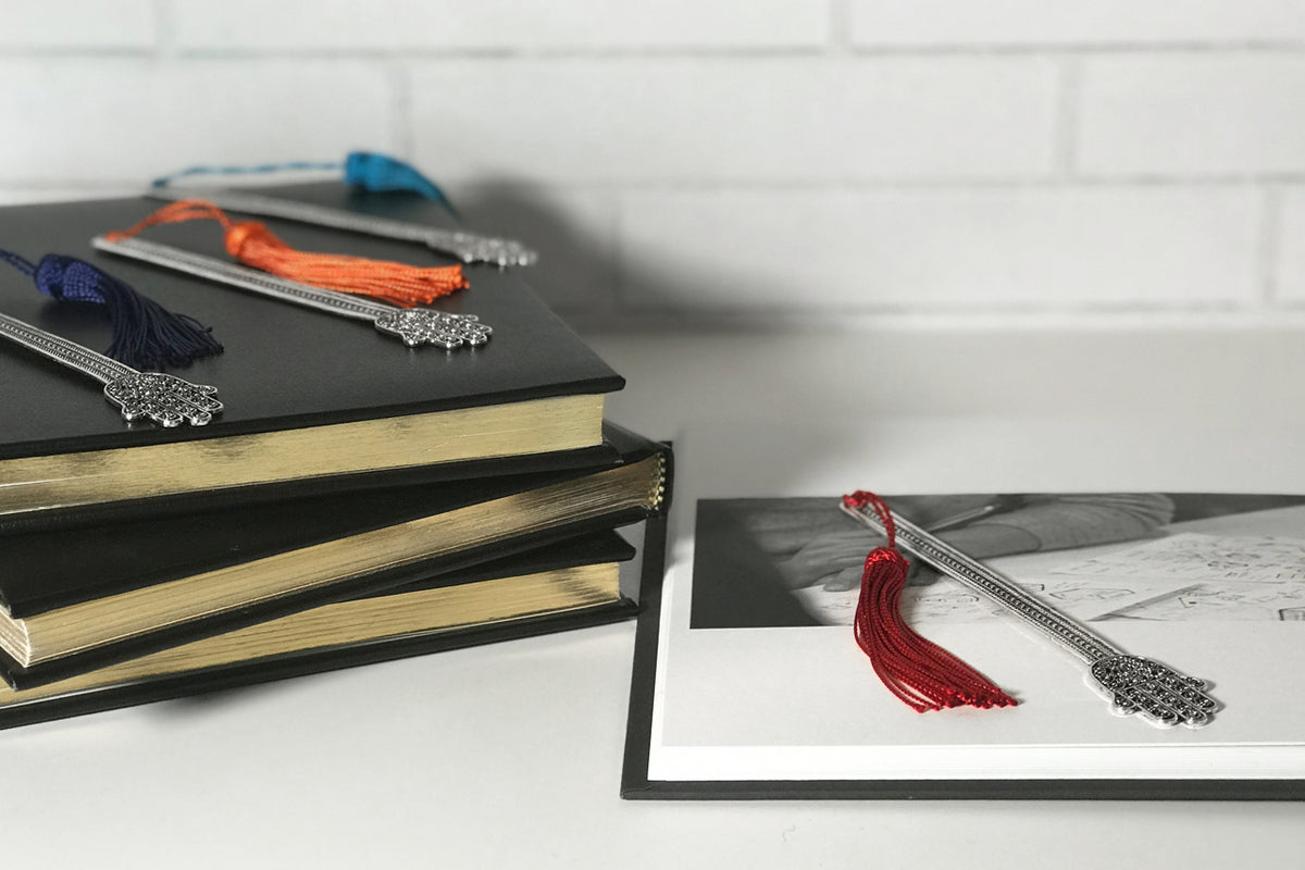 Hand of Fatima bookmarks with ornate metal charms and colored tassels displayed on stacked books and an open page.