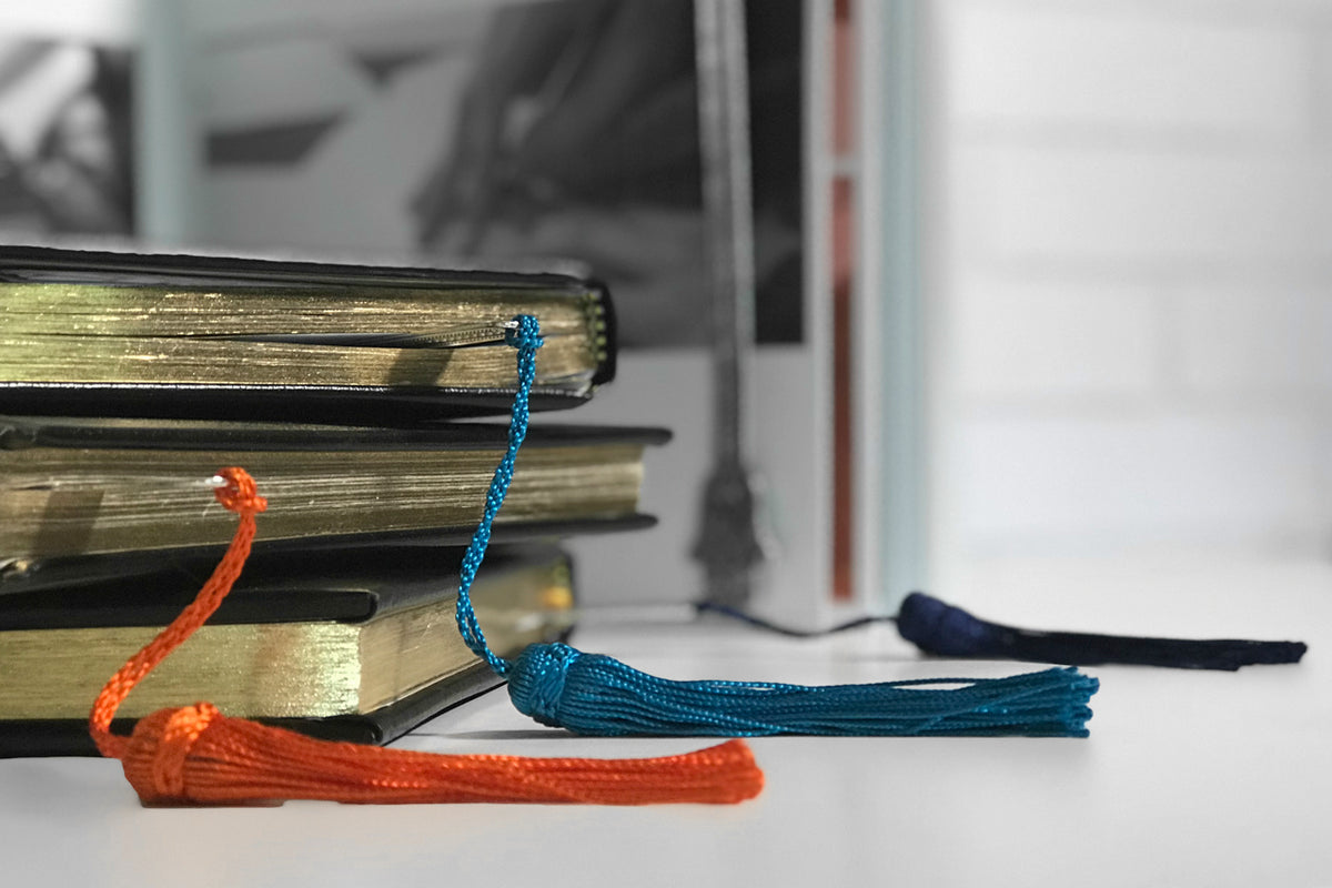 Bookmark with hand-shaped metal charm and braided cord tassel in orange and blue, displayed with stacked books.