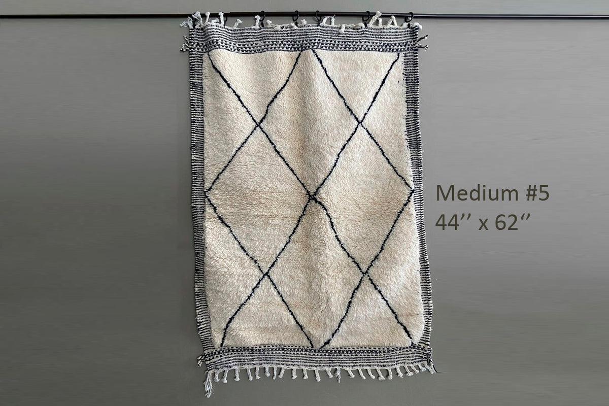 Cream Berber rug with black diamond pattern, fringed edges, and textured border on gray background.