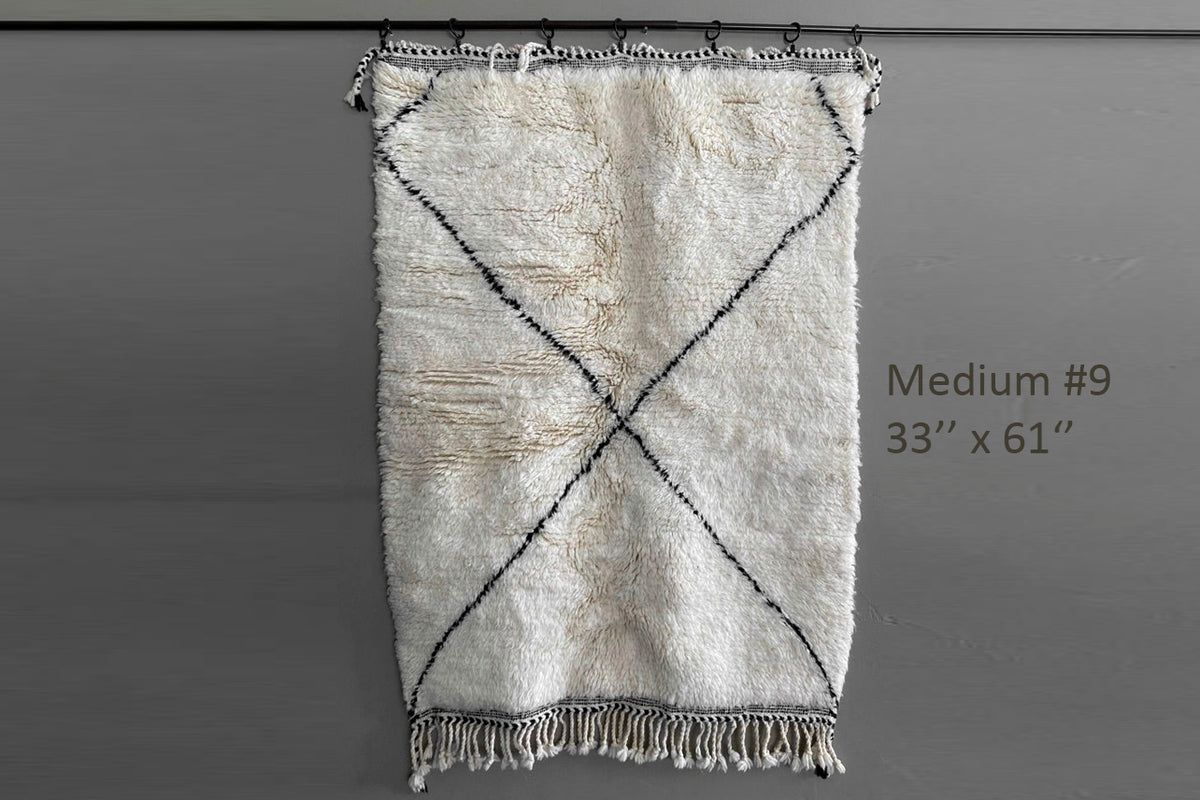 Cream-colored Berber rug with dark diamond cross pattern, fringe edges, and geometric designs, displayed flat.