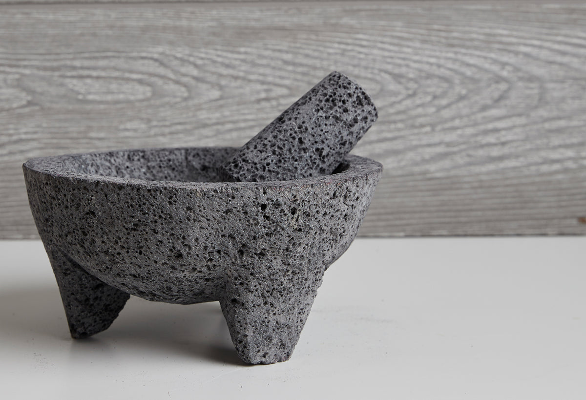 Gray stone molcajete with cylindrical pestle resting inside the textured mortar bowl on white surface.