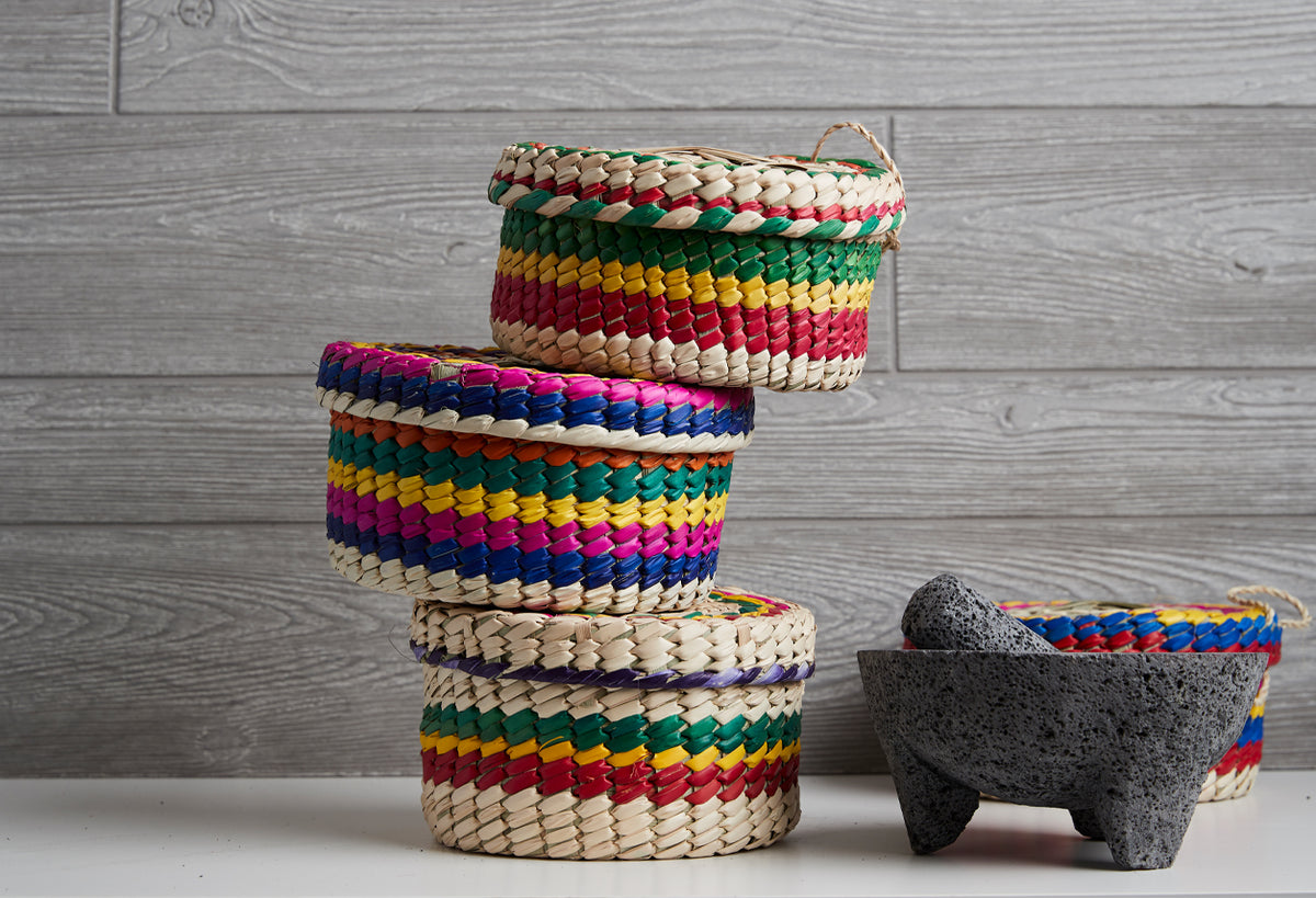 Stacked woven baskets with colorful fabric trim and dark stone molcajete with pestle on white surface.
