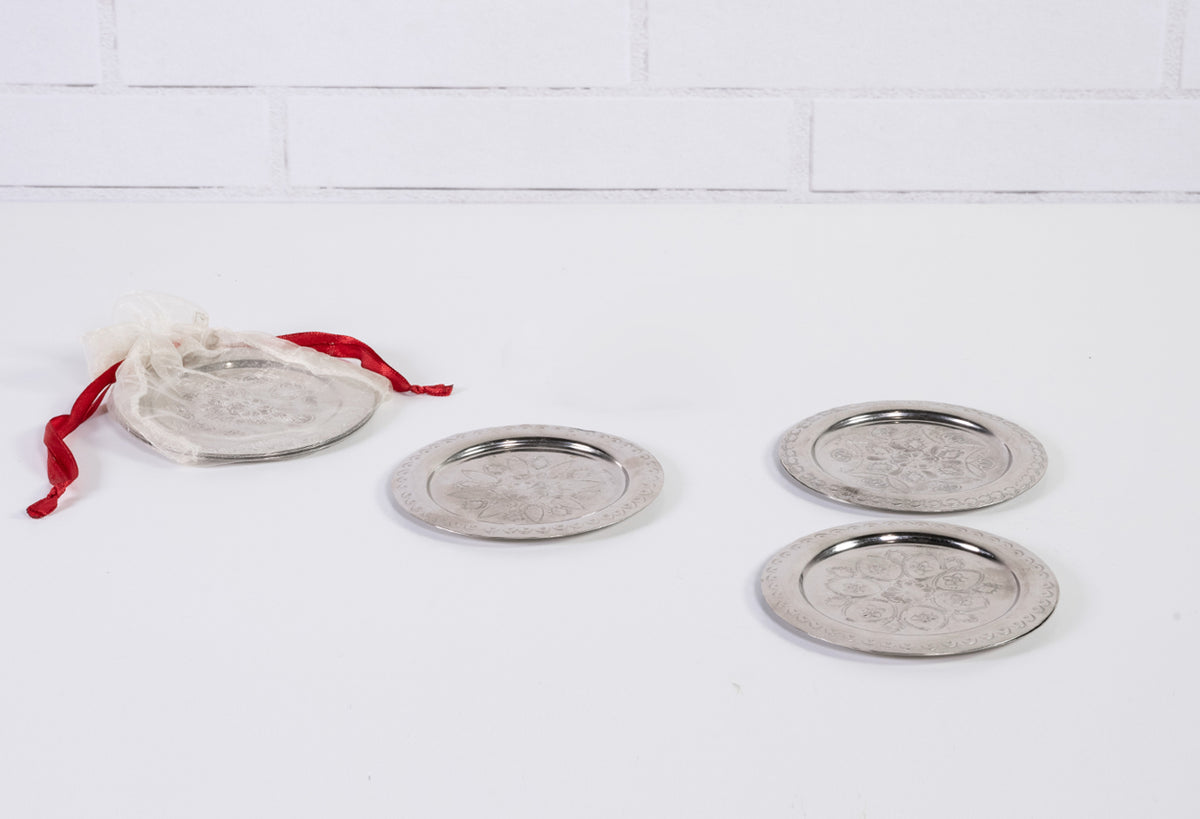 Four round silver coasters with etched geometric patterns and metal rims, one with red fabric drawstring bag.