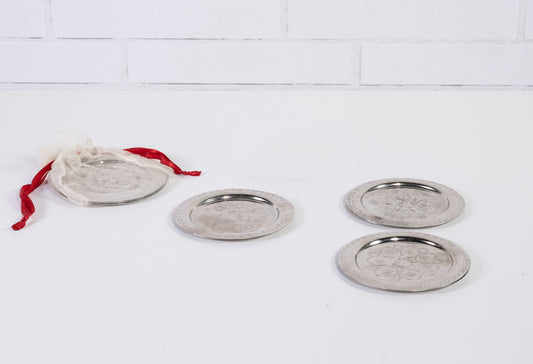 Four round silver coasters with etched geometric patterns and metal rims, one with red fabric drawstring bag.