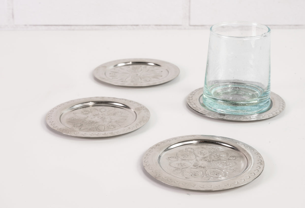 Four silver-toned round coasters with etched geometric patterns, one holding a clear glass.