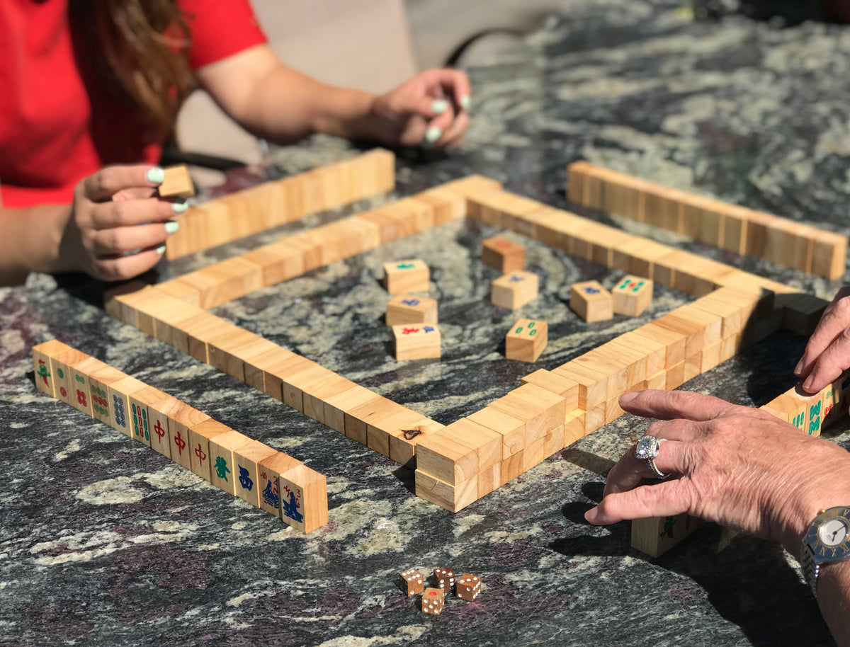 Mahjong game set with wooden tiles and rack being played by two people on a gray patterned surface.
