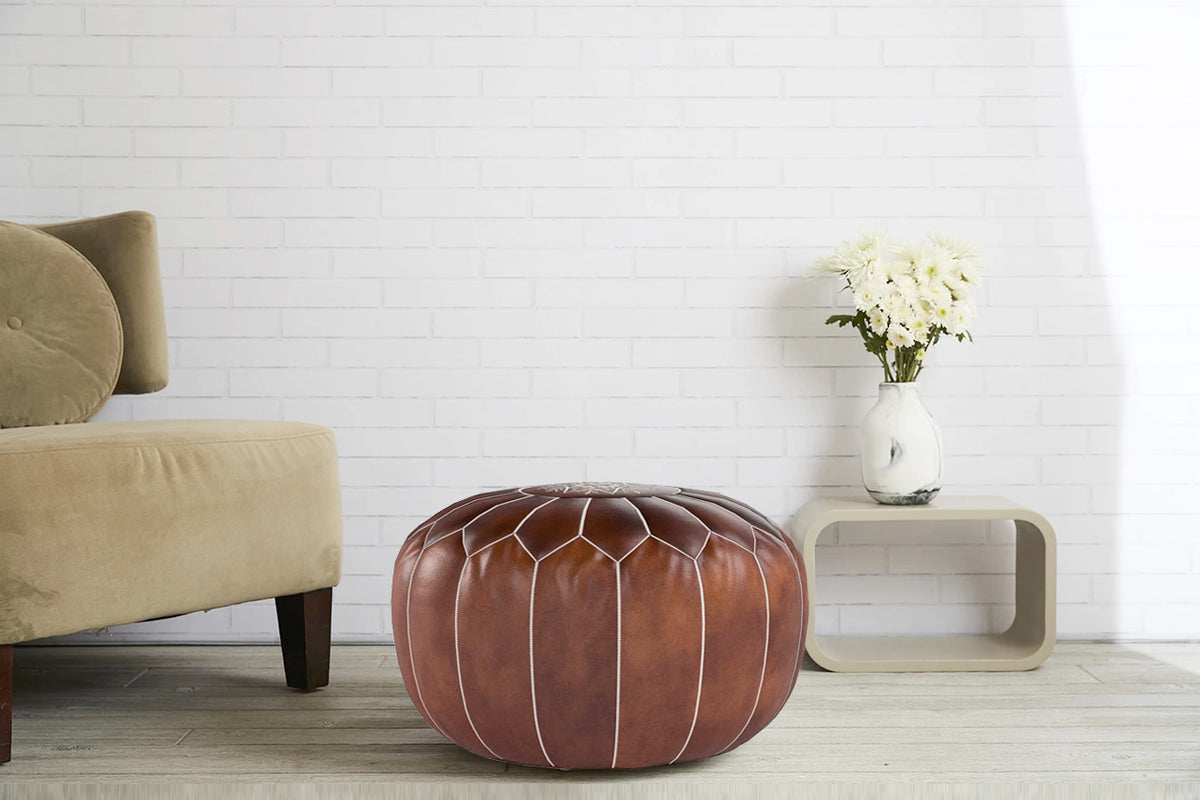 Brown leather pouf with white stitching detail, styled next to a beige chair in a living room setting.