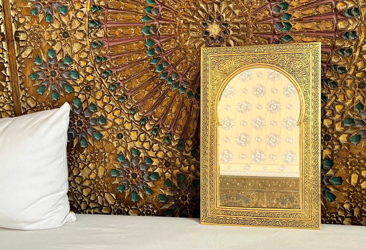 Ornate gold-framed mirror with arched top and decorative border displayed against patterned gold and teal backdrop.