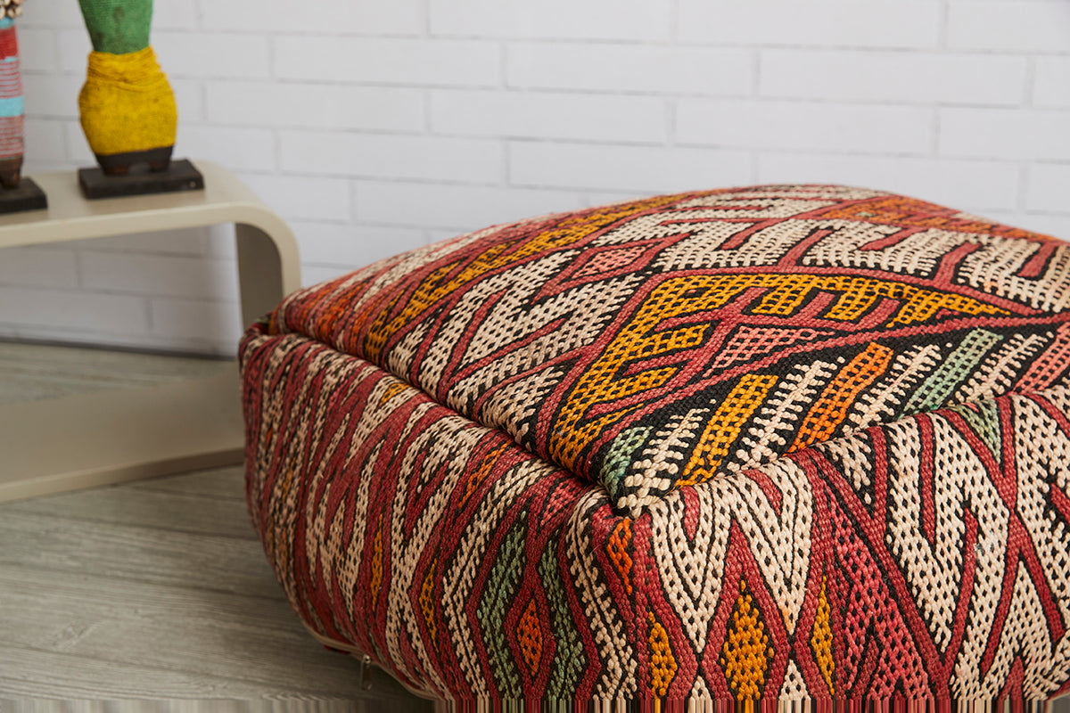 Moroccan kilim pouf with geometric pattern in red, yellow, cream, and green stripes on woven fabric.