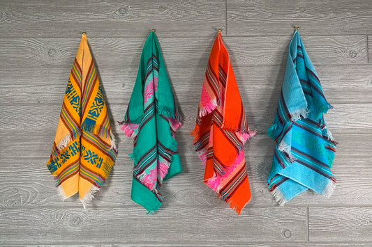 Four folded cloth napkins in yellow, green, orange, and blue with geometric patterns and pink tassels.