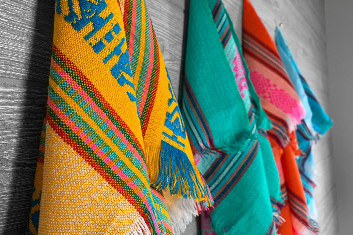 Multiple colorful patterned napkins with geometric designs and fringed edges, featuring yellow, teal, red, and blue colors.
