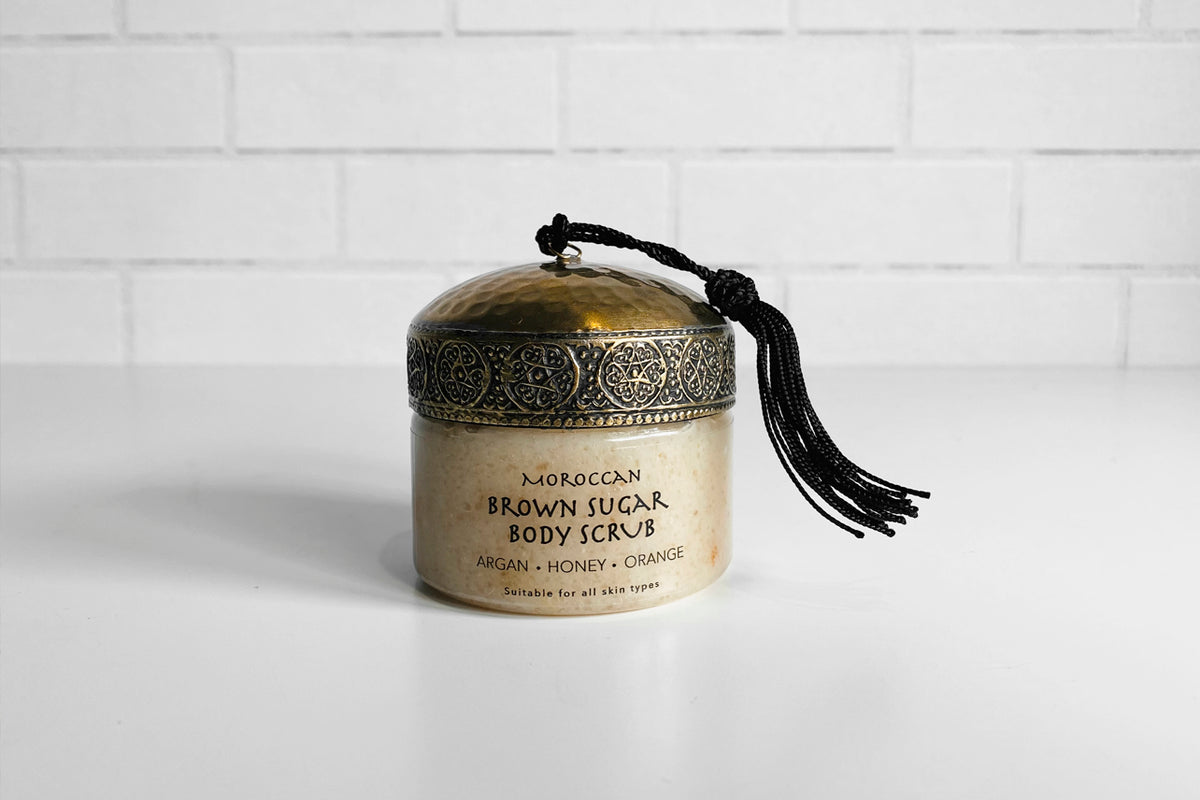 Cream-colored body scrub jar with decorative metallic lid and black tassel, labeled Moroccan Brown Sugar Body Scrub.