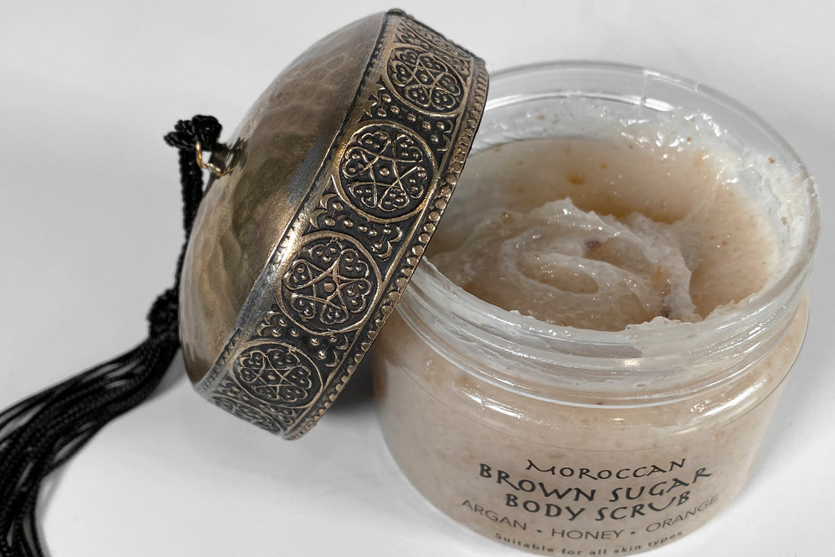 Moroccan Brown Sugar Body Scrub in clear glass jar with ornate metal lid and black tassel.