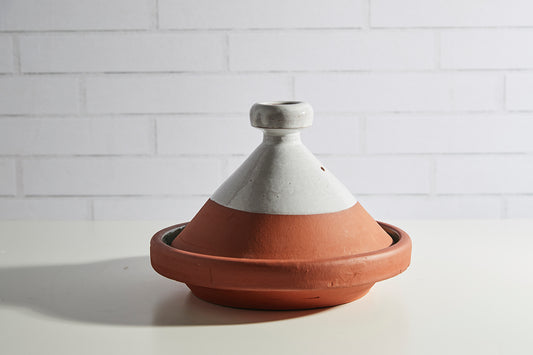 Terracotta tagine with cream-colored conical lid and two handles on white brick background.
