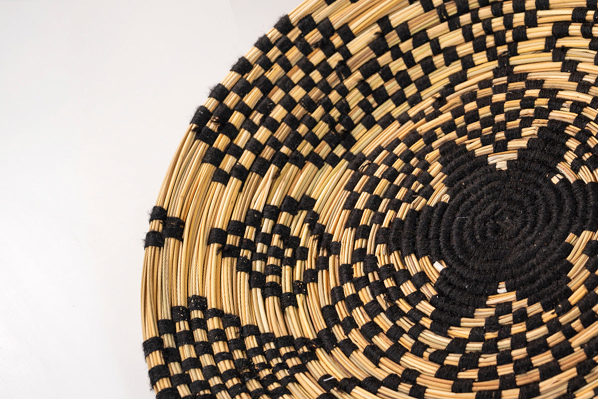 Round woven tray with natural straw and black geometric diamond pattern design.