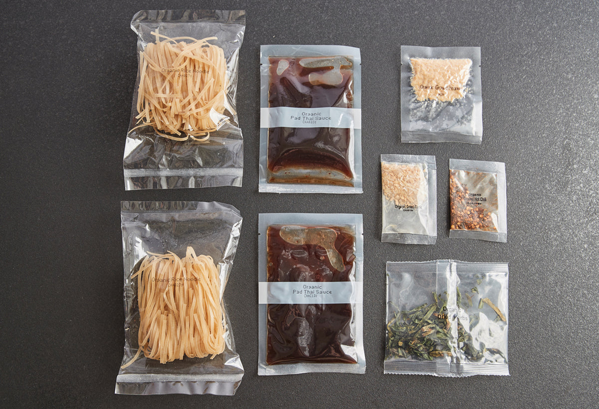 Meal kit containing two packages of dried noodles, two sauce packets, and five smaller packets of ingredients arranged on gra