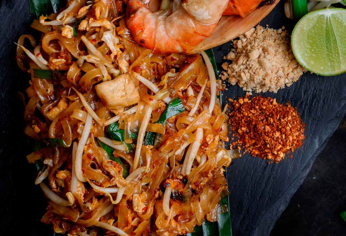 Pad Thai noodles with shrimp and chicken on dark slate with lime, peanuts, and spice powder.