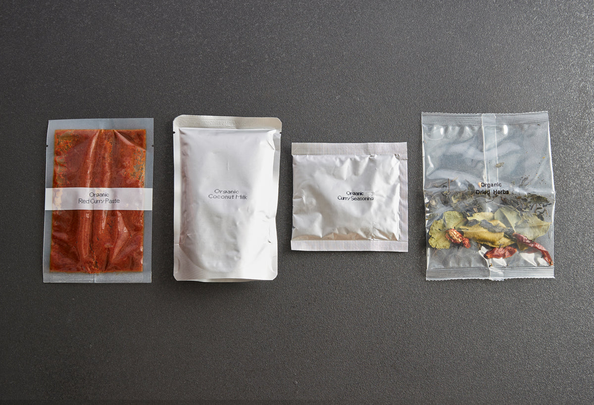 Four sealed packets containing red curry paste, coconut milk, curry sauce, and dried herbs arranged on gray surface.