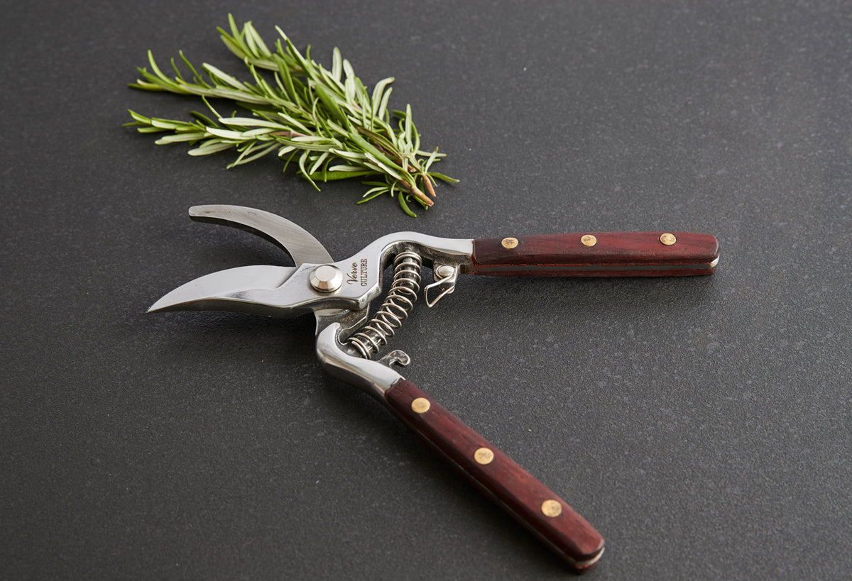 Curved pruning shears with dark wood handles and brass rivets next to a sprig of fresh rosemary on gray surface.