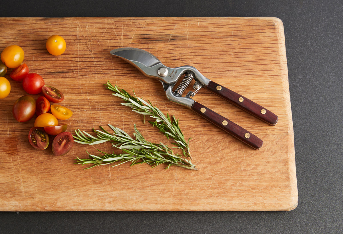 Pruning shears with dark wood handles and metal blades on wooden cutting board with tomatoes and rosemary sprigs.