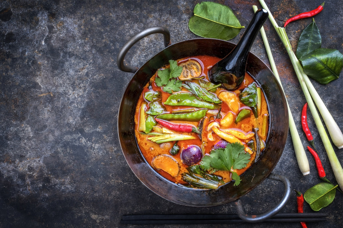 Black wok with Thai curry containing vegetables, chopsticks, fresh herbs, and red chilies on dark surface.