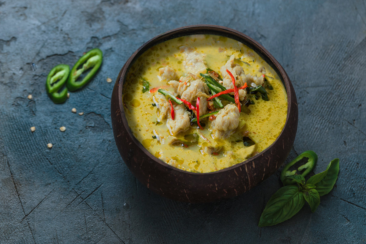 Yellow curry soup with chicken, red chilies, and basil in a brown bowl on blue surface with sliced green chilies nearby.
