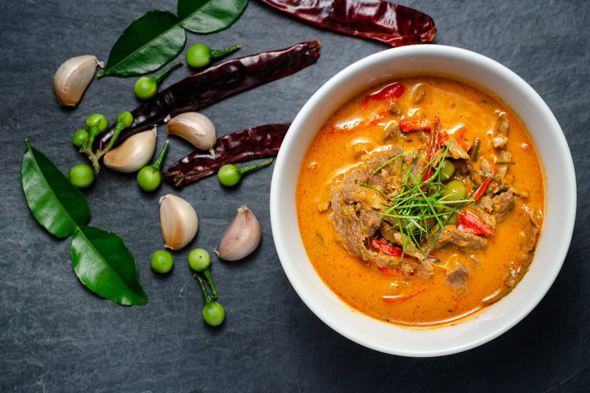 White bowl of orange curry with meat and herbs, surrounded by garlic, green peppercorns, chilies, and fresh herbs on gray sur
