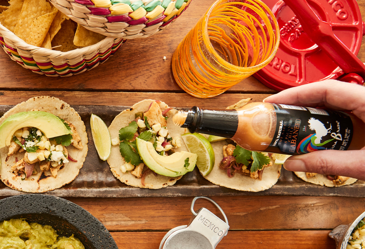 Habanero hot sauce bottle being drizzled over tacos topped with avocado, meat, and cilantro.